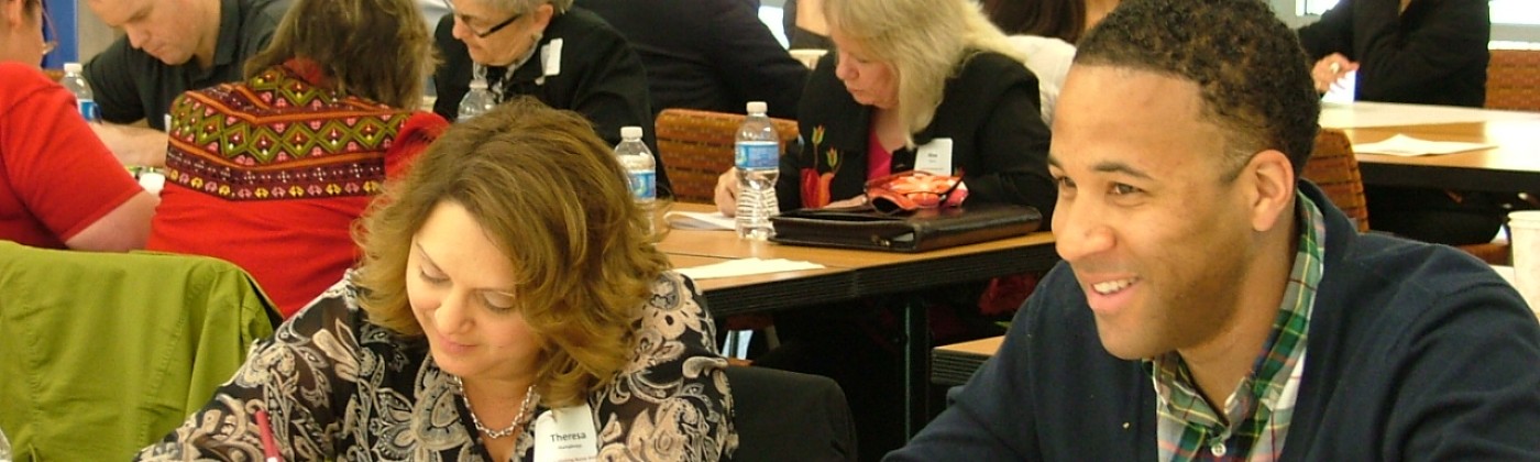 A woman and a man working side-by-side at a table, in a room full other other people working at tables.