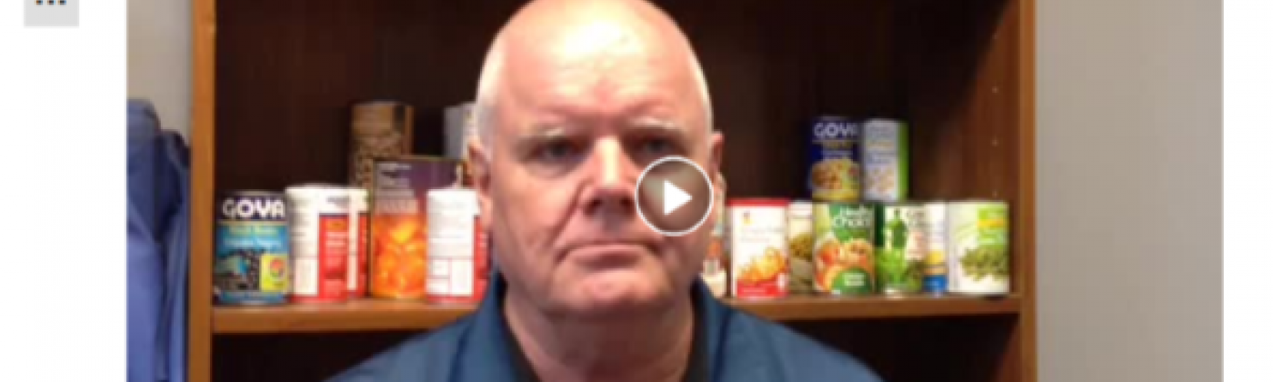 Still of a video of a Caucasian man in front of shelves with canned goods.
