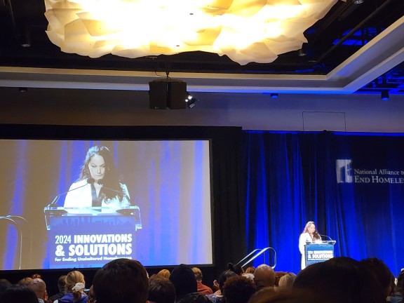 National Alliance to End Homelessness' CEO Ann Olivia is speaking at a podium on a blue-lit stage to conference attendees during the opening plenary..