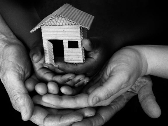 Image of three people's hands laid on top of each other, cradling a small wooden house