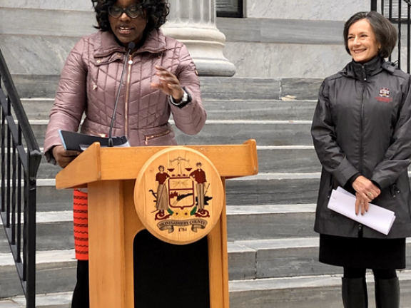 Migrated automatically. Director of Health & Human Services speaking at podium with Commissioner smiling