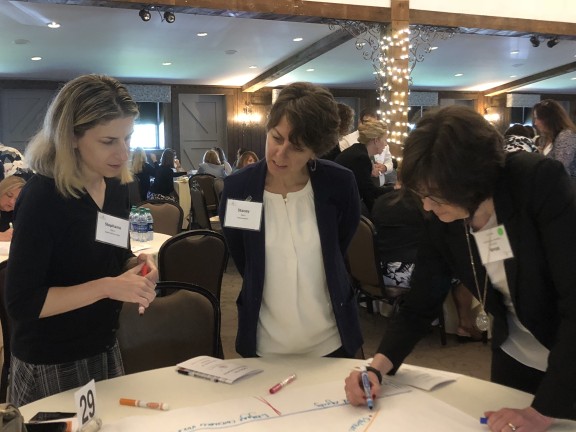 Three women looking down at a large sheet of paper on a table; one of the women is writing on the sheet.