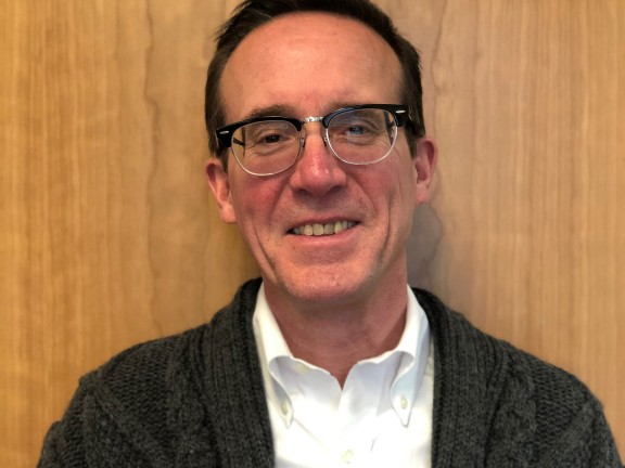 A man in front of a wood panel, in glasses, a white shirt and gray knit sweater