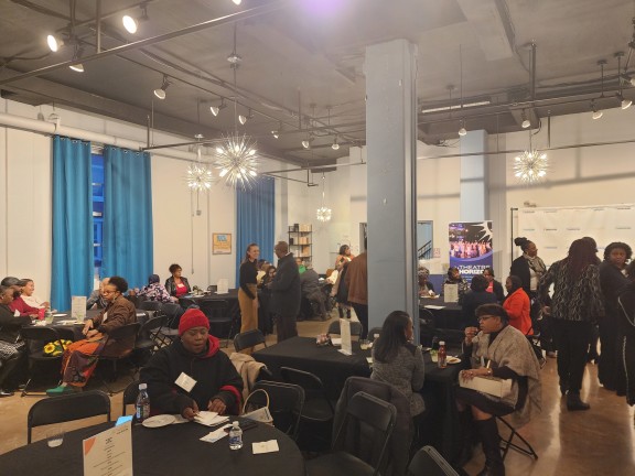 A group of people standing and sitting at tables, talking to one another. 