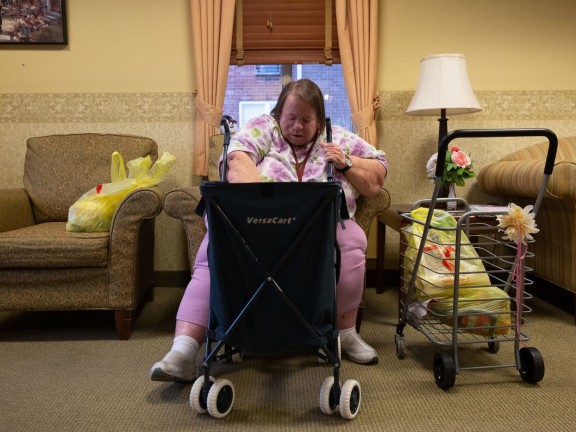 An elderly woman sits and digs around in a shopping cart in front of her.