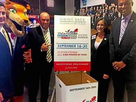 Migrated automatically. Four people and a person in a mascot costume, standing by a box for food donations.