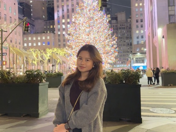 A picture of Ha Nguyen, our intern. Ha is Vietnamese, with light brown long hair. She's standing in front of a lit Christmas tree in NYC and is wearing a black sweater and a gray coat.