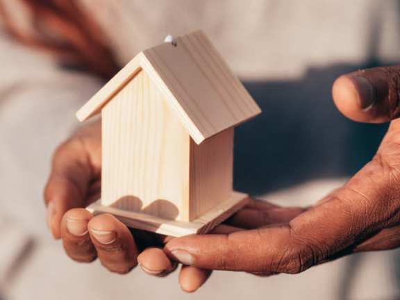 A picture of a couple with their hands holding a miniature wooden house together.