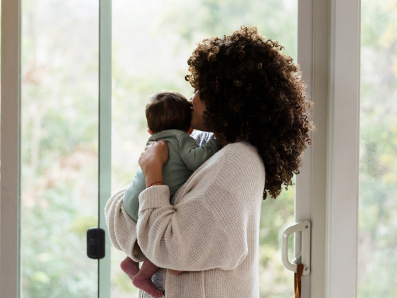A picture of a mom holding her baby, as they look at out a window.