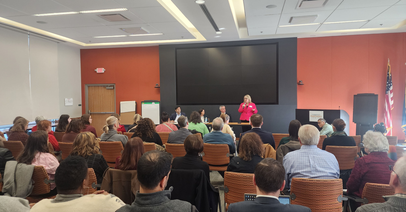 A lady wearing a pink blazer, holding a microphone, introducing three speakers, sitting at a table next to her.