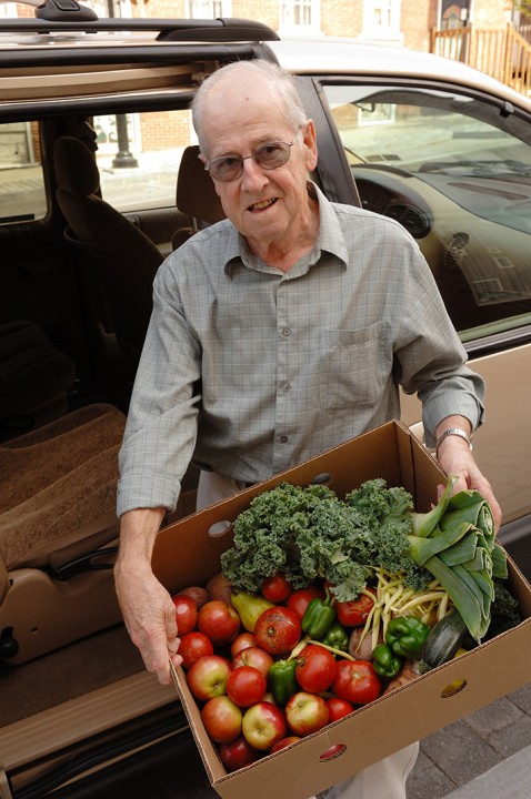 An elderly man next to a car, holding a cardboard box with fresh produce