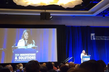 National Alliance to End Homelessness' CEO Ann Olivia is speaking at a podium on a blue-lit stage to conference attendees during the opening plenary..
