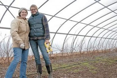 Two women bundled up in coats in a hoop house, standing over tilled soil.
