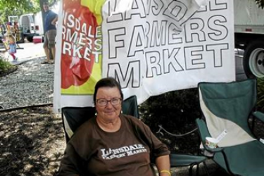 A woman sitting in a lawn chair in front of a banner that reads, "Lansdale Farmers Market."