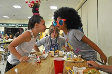 Two girls staring at each other over a school lunch table with food, a boy looks at both of them.