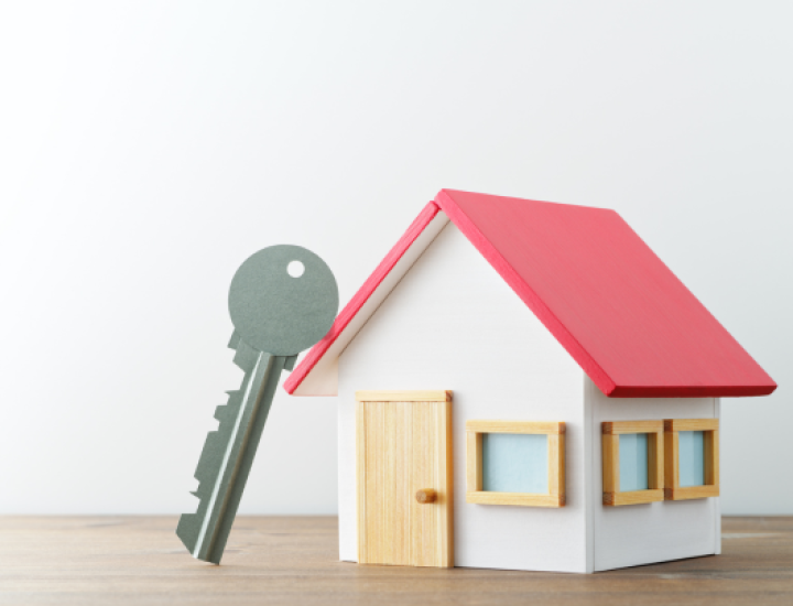 A picture of a toy house with a red roof, three windows, a door, with a silver key leaning against it.